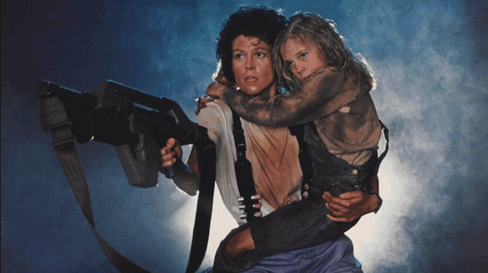 Film still of a woman holding a young girl on one arm and a large gun in the other hand