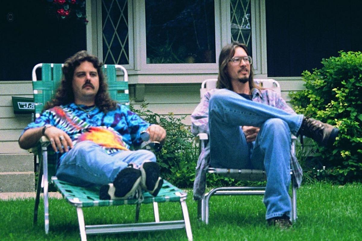 Film still of two men with long hair sitting in lawn chairs.