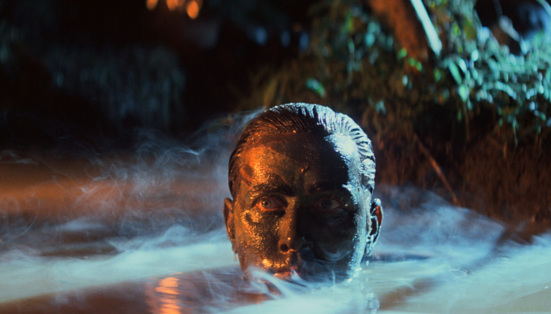 Film still of a man (Martin Sheen) with his face covered in mud and only the top half of his head sticking out of water.