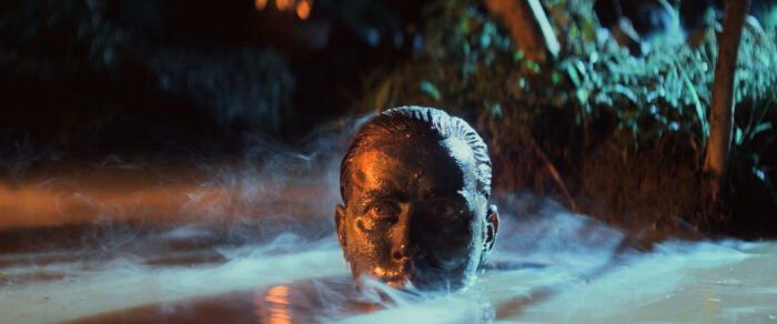 Film still of a man (Martin Sheen) with his face covered in mud and only the top half of his head sticking out of water.