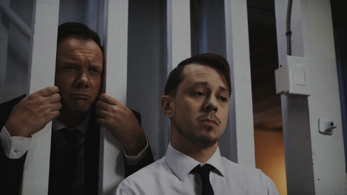 Film still of two men in suits looking upset. One is sticking his face in between two white bars.