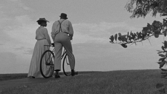 Black and white film still of a man and woman in early 20th century formal clothes walking up a hill. One of them is pushing a bike.