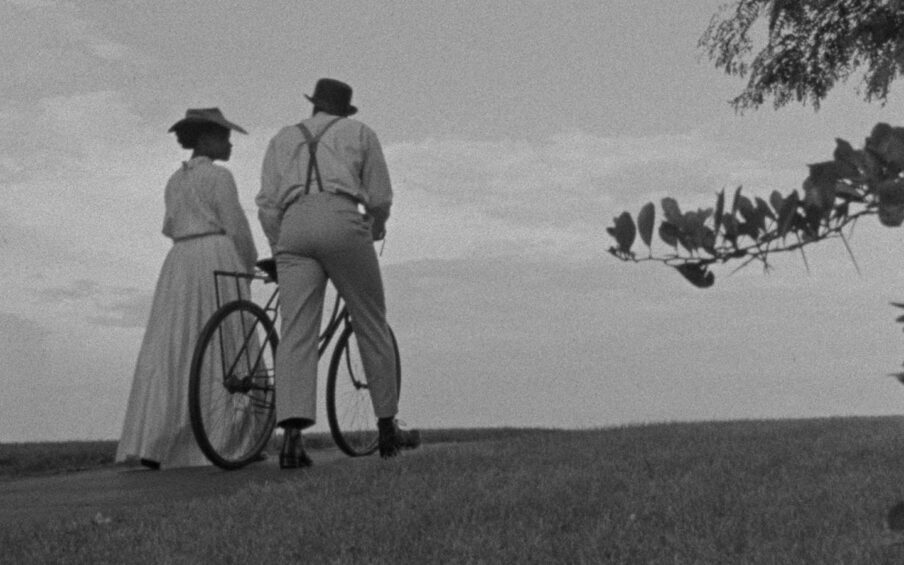 Black and white film still of a man and woman in early 20th century formal clothes walking up a hill. One of them is pushing a bike.