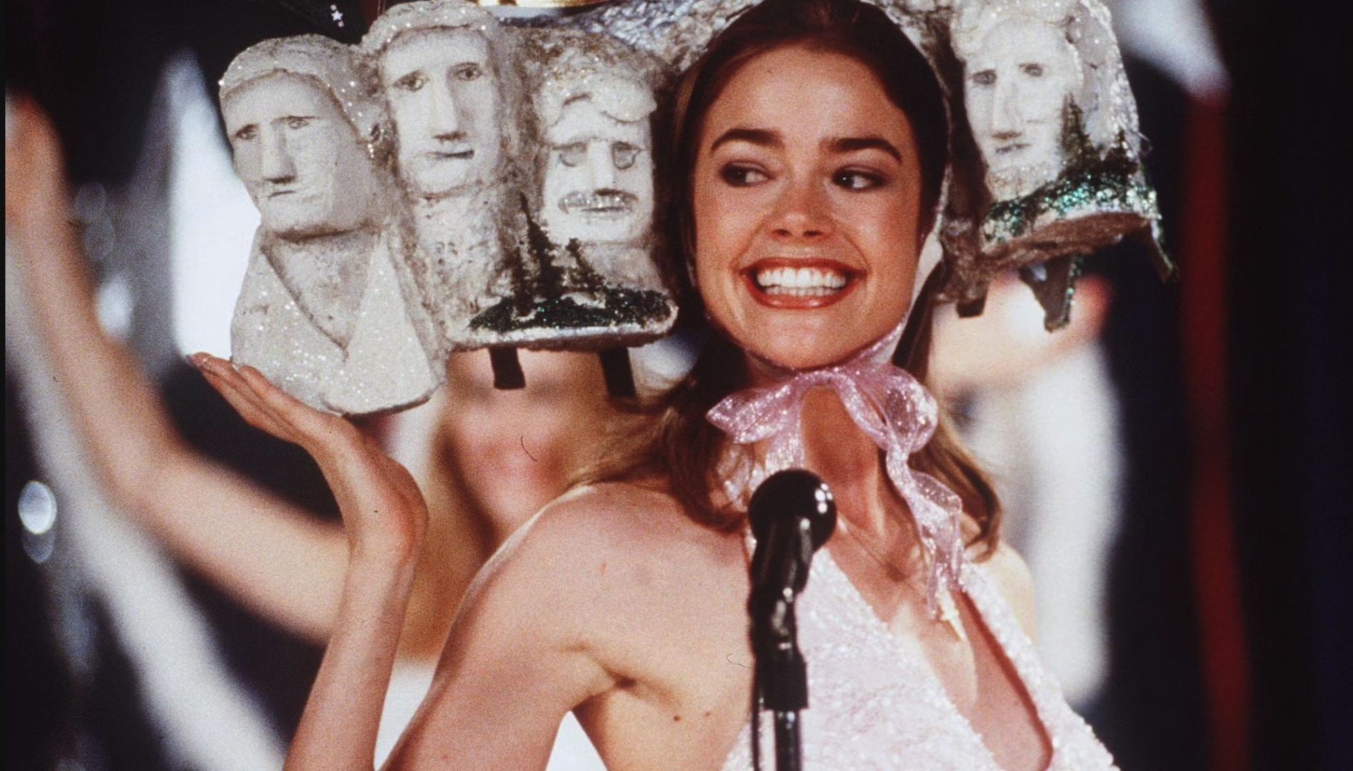 Photo of a smiling woman at a microphone, wearing a model of Mount Rushmore on her head.