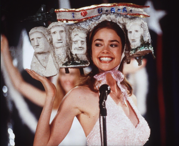 Photo of a smiling woman at a microphone, wearing a model of Mount Rushmore on her head.
