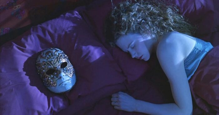 Film still of a woman with curly red hair sleeping. On the pillow next to her is an ornate mask.