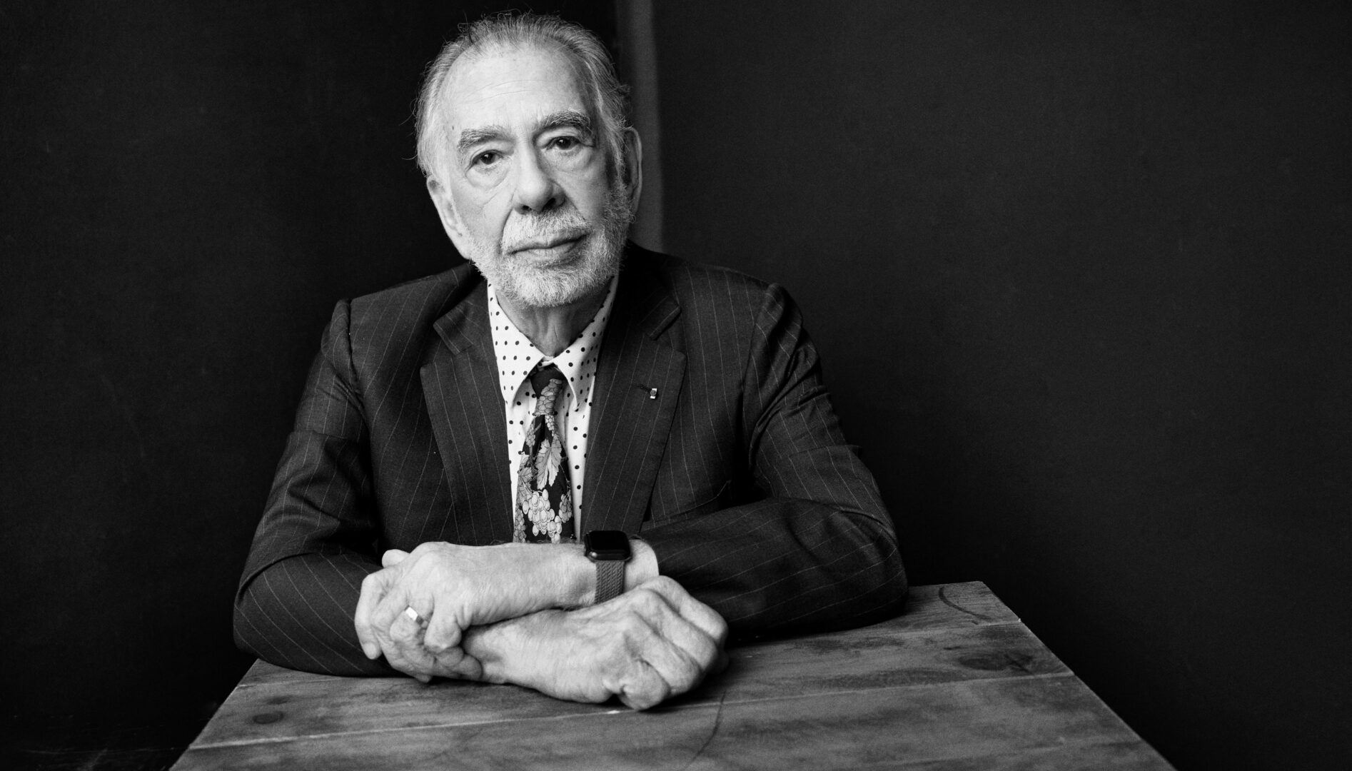 Black and white portrait of a man (Francis Ford Coppola) with white hair, wearing a suit and tie, sitting at a table.
