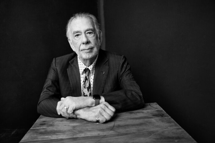 Black and white portrait of a man (Francis Ford Coppola) with white hair, wearing a suit and tie, sitting at a table.