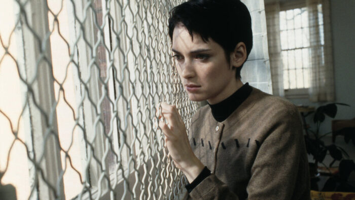 Photo of a young woman with short dark hair standing in front of a chainlink-covered window.