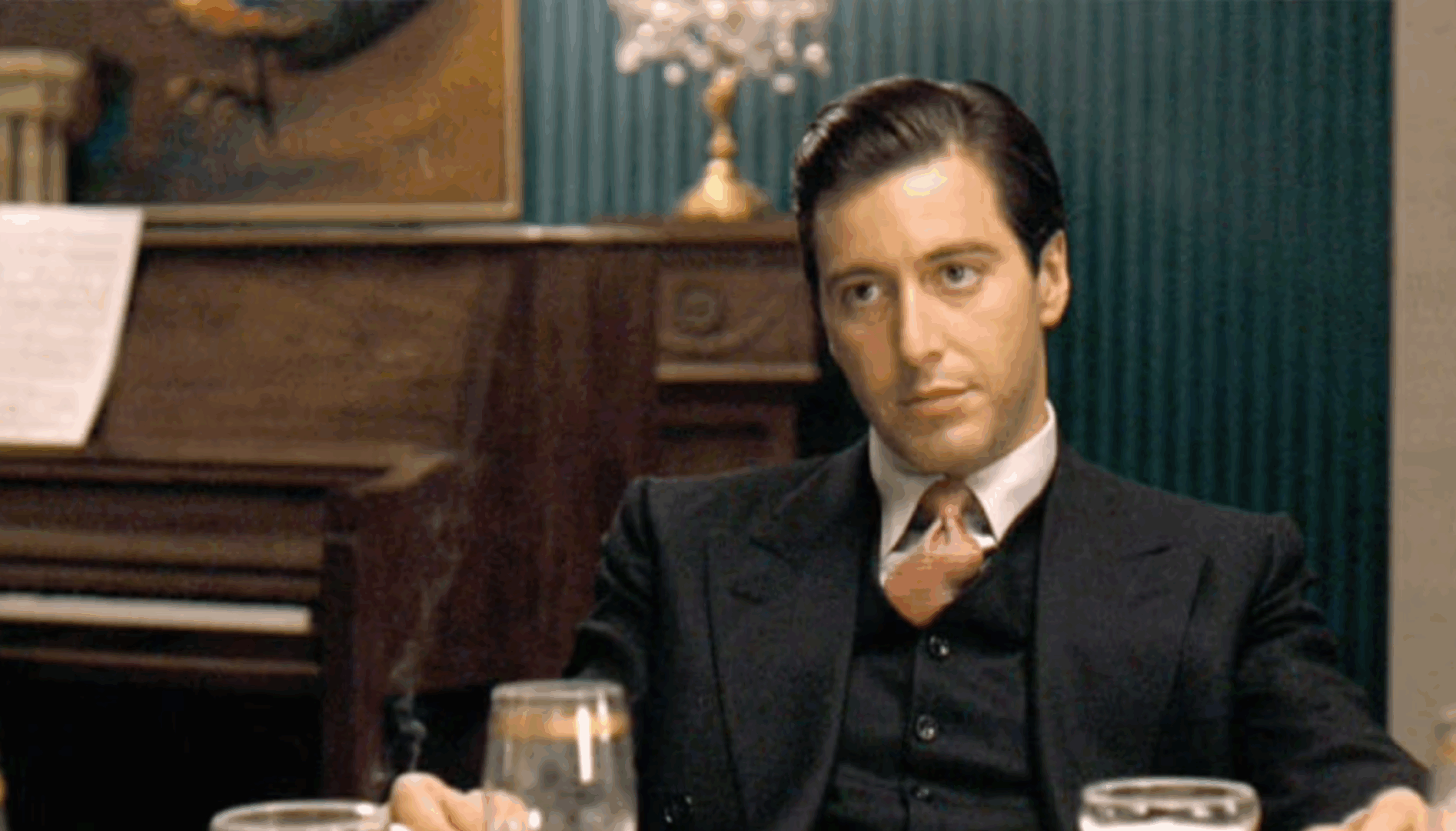 Film still of a young man (Al Pacino) with short dark hair, wearing a suit, sitting at a table with a piano behind him.