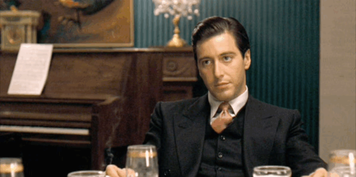 Film still of a young man (Al Pacino) with short dark hair, wearing a suit, sitting at a table with a piano behind him.