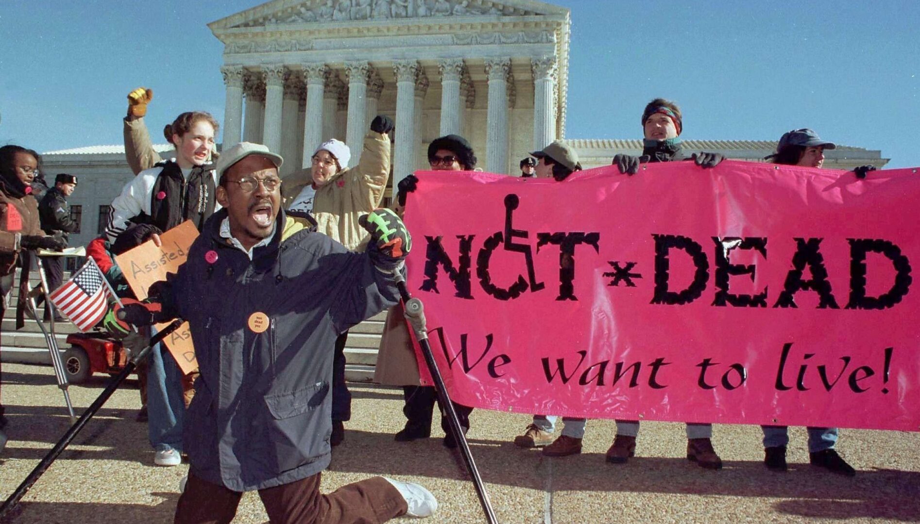 Film still of a group of protestors outside the Supreme Court building. A group are holding a pink sign that says "Not Dead / We Want to Live"