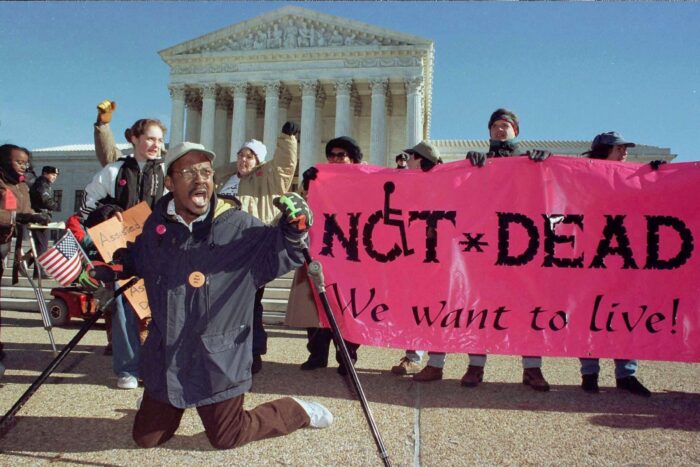 Film still of a group of protestors outside the Supreme Court building. A group are holding a pink sign that says "Not Dead / We Want to Live"