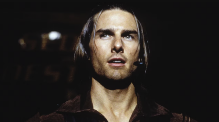 A film still of a white man with long brown hair (Tom Cruise) wearing a wireless microphone.