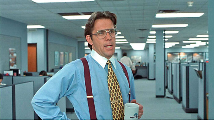 Film still of a man in a blue button down shirt, tie, and suspenders, holding a coffee cup in a row of cubicles.
