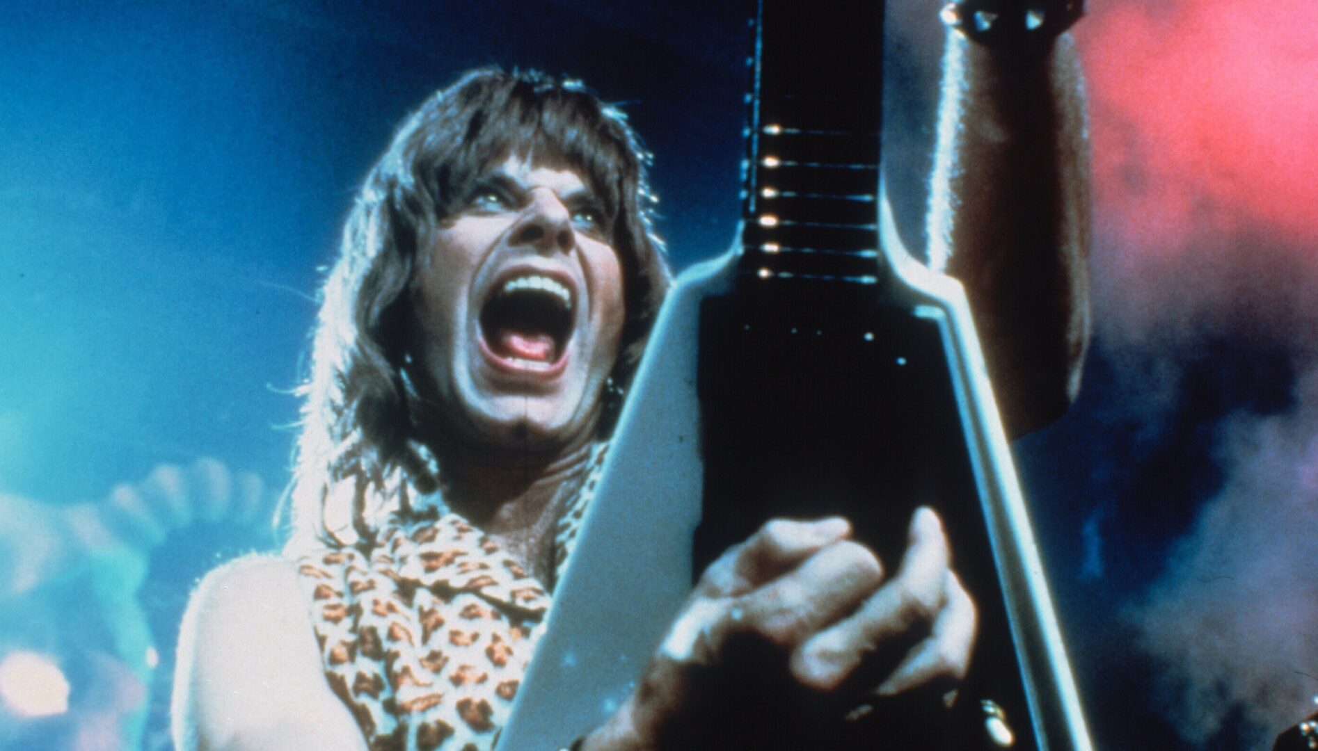 Film still from This is Spinal Tap. Image of a man on stage holding a guitar up with his mouth open. Lights and smoke are behind him.