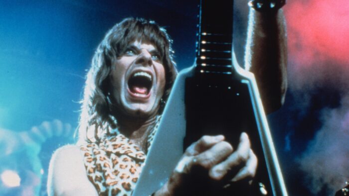 Film still from This is Spinal Tap. Image of a man on stage holding a guitar up with his mouth open. Lights and smoke are behind him.