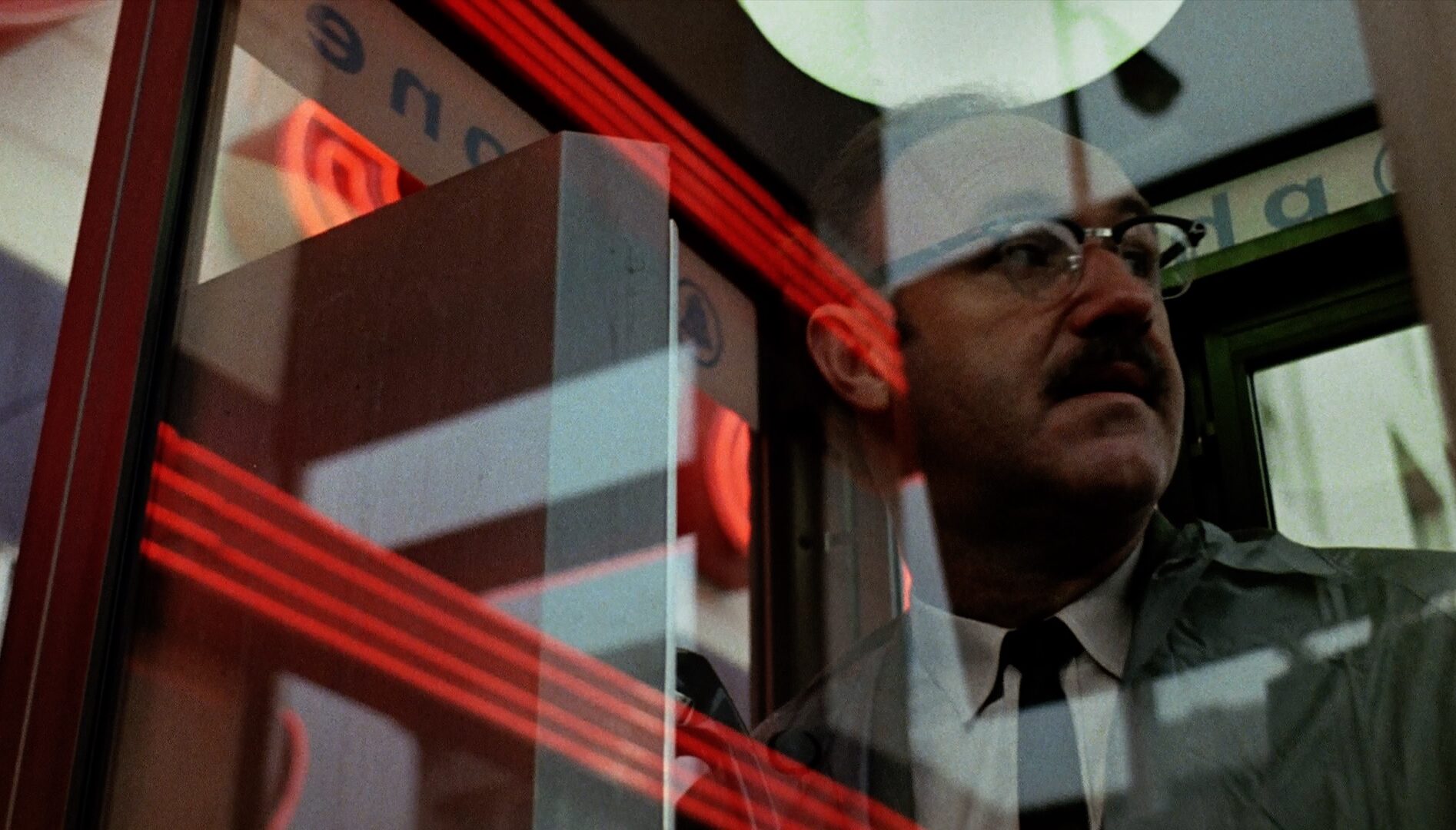 Film still of a man with eyeglasses (Gene Hackman) behind a pane of glass with reflections of red lights on it.