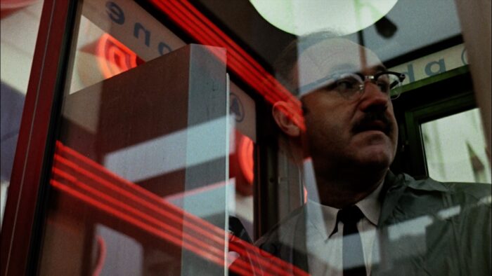 Film still of a man with eyeglasses (Gene Hackman) behind a pane of glass with reflections of red lights on it.