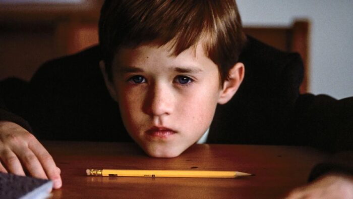 Film still of a young boy resting his chin on a desk. A pencil is on the desk in front of him.