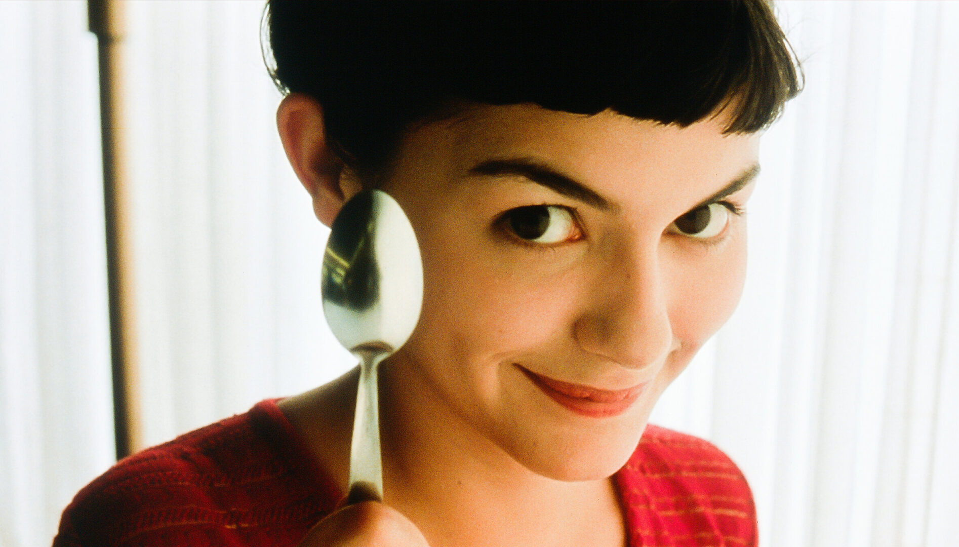 Film still close-up of a young woman with short dark hair, holding up a spoon to the camera and smiling.