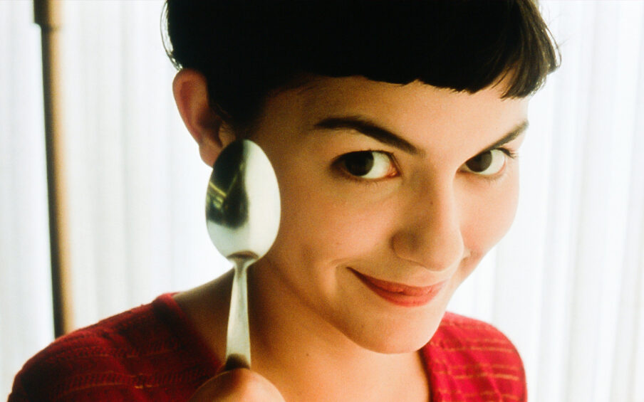 Film still close-up of a young woman with short dark hair, holding up a spoon to the camera and smiling.