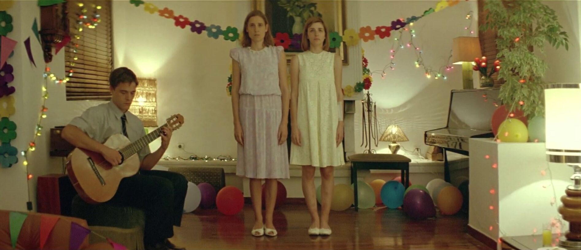 Film still of a dimly lit room where a young man sits playing guitar on the left and two young women in dresses stand in the middle in front of decorations.