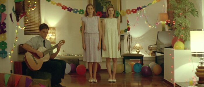 Film still of a dimly lit room where a young man sits playing guitar on the left and two young women in dresses stand in the middle in front of decorations.