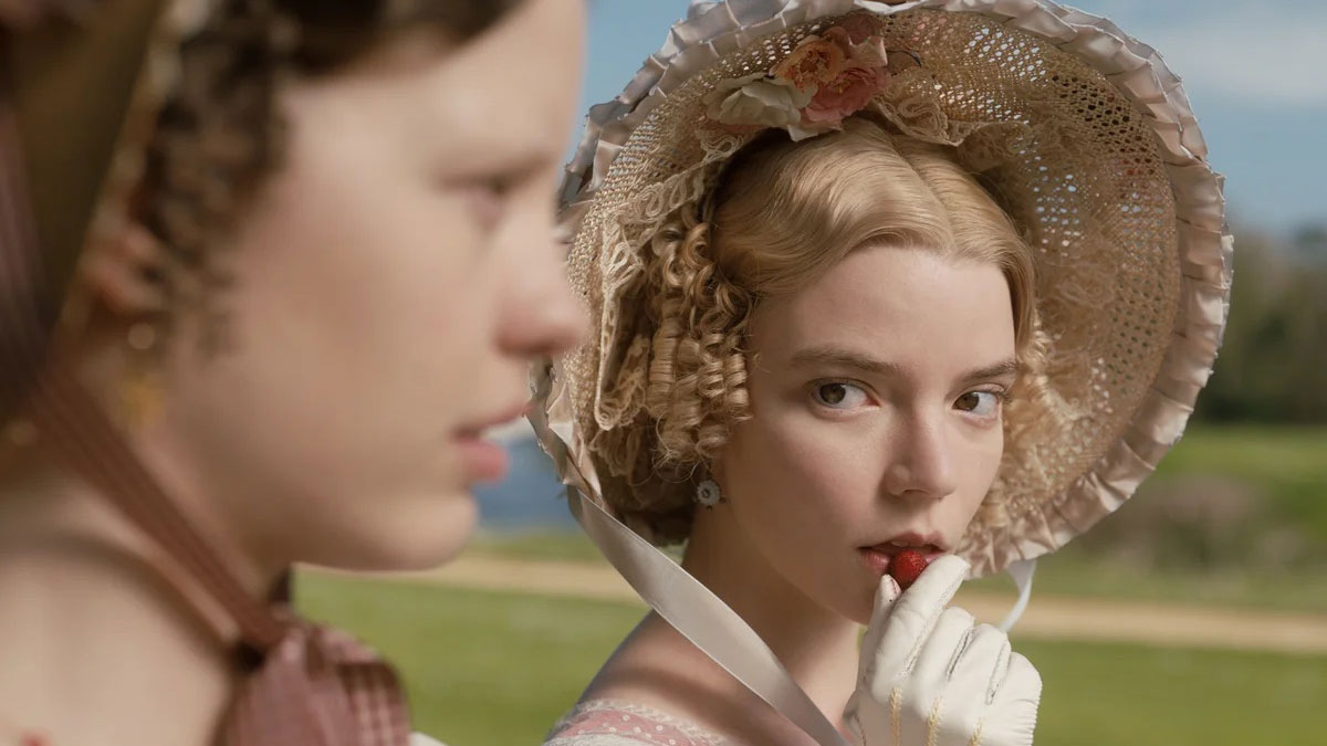 Film still of a woman with blonde curly hair, wearing a 19th century bonnet with flowers on it, and holding a berry up to her mouth with a white-gloved hand. She is staring at the side of another young woman's face.