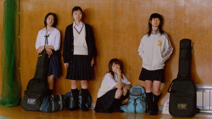 A group of young women in school uniforms, standing against a wood-paneled wall. They have backpacks and instruments in cases with them.