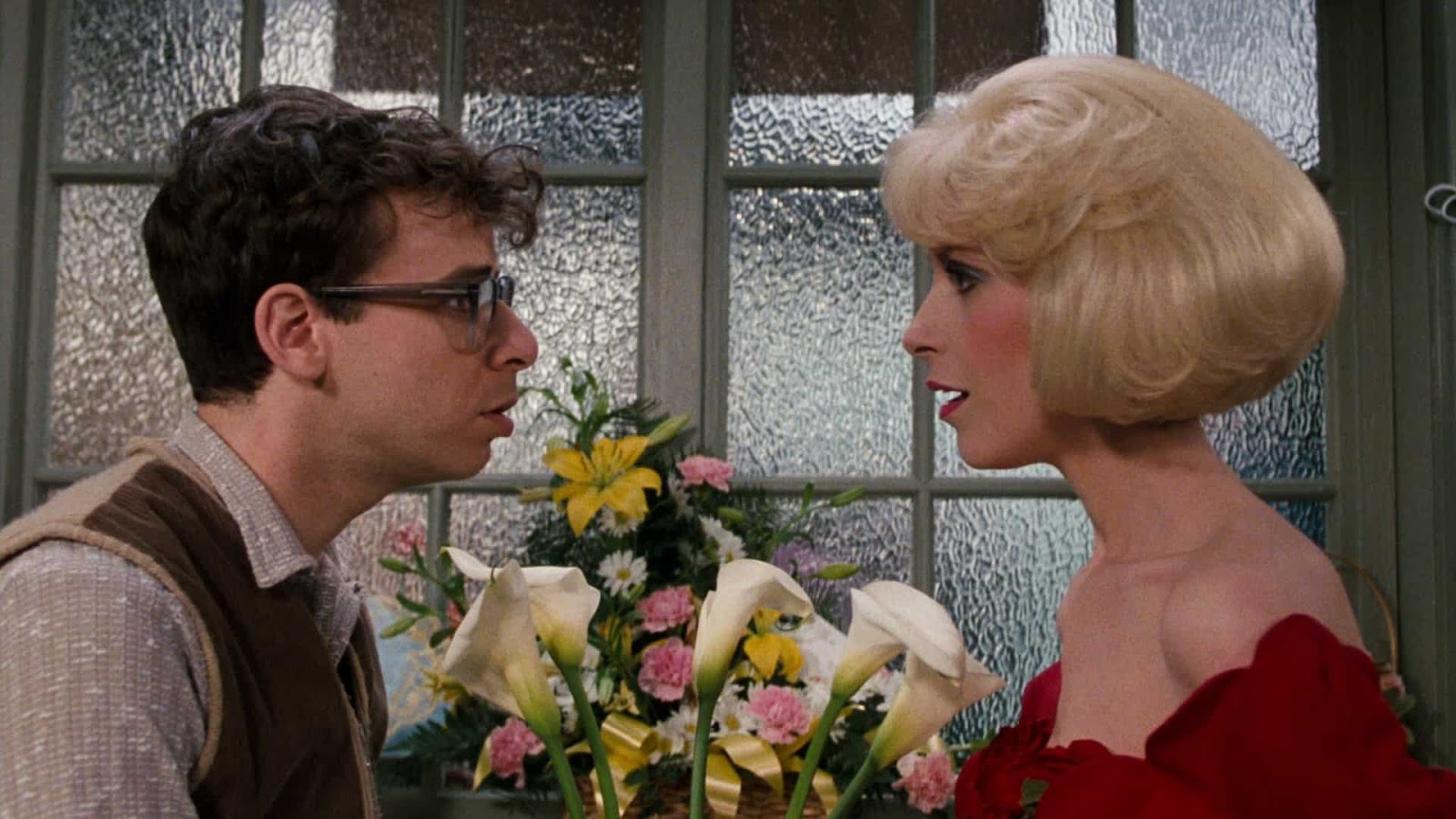 Film still of a man with short brown hair and eyeglasses looking at a woman with big blonde hair. A large bouquet of flowers is between them.