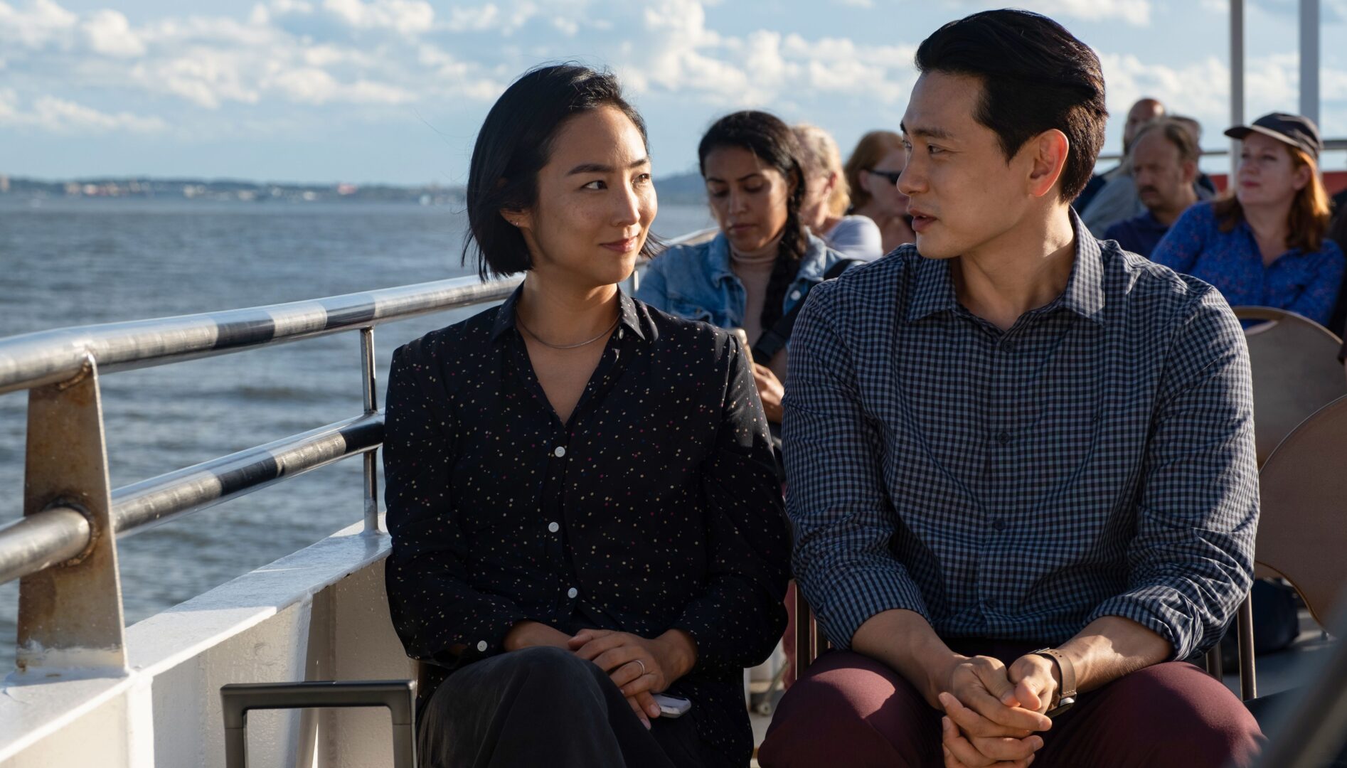 A Korean man and woman sitting next to each other on the top deck of a ferry boat. They are looking at each other, her with a smile on her face.