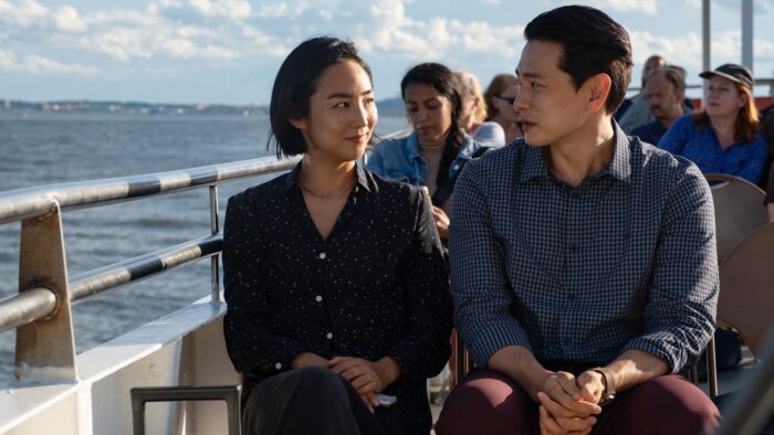 A Korean man and woman sitting next to each other on the top deck of a ferry boat. They are looking at each other, her with a smile on her face.