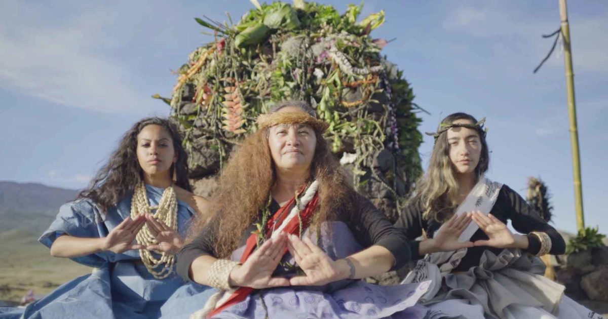 Film still of three woman with long hair and Native Hawaiin outfits sitting outside in front of a large plant arrangement.