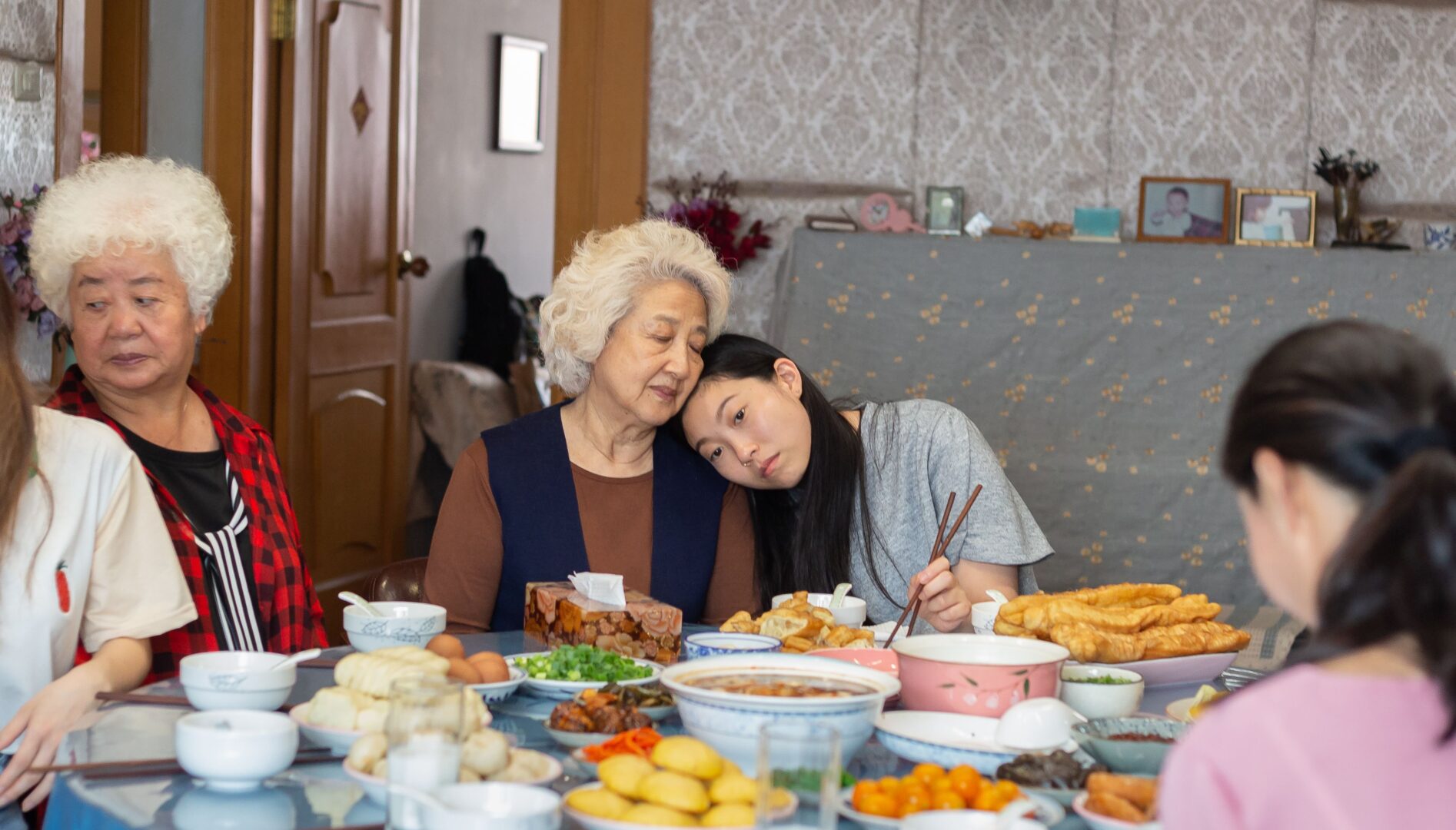Film still of a restaurant table covered in dishes of food. A multi-generational family sits around it. In the foreground is a white-haired older woman resting her head on a young brown-haired woman sitting next to her.