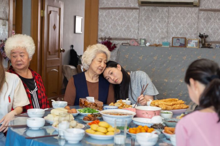 Film still of a restaurant table covered in dishes of food. A multi-generational family sits around it. In the foreground is a white-haired older woman resting her head on a young brown-haired woman sitting next to her.