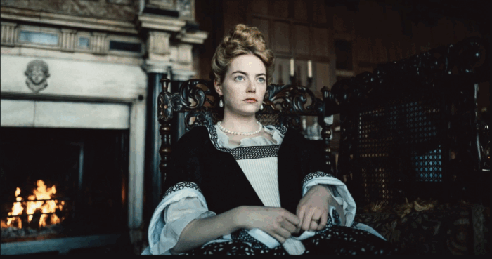 Film still of a pale woman with curly blonde hair and a formal 19th century dress, sitting in front of a fireplace.