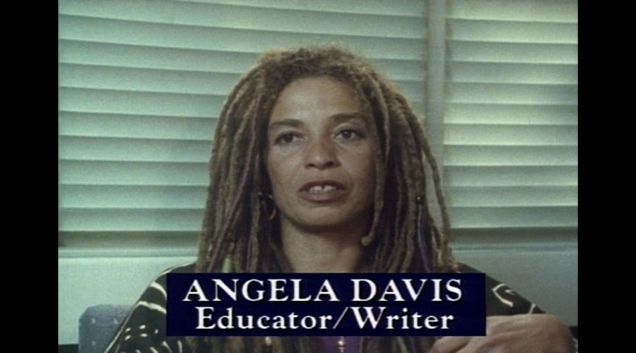 Film still of a Black woman (Angela Davis) with long dreadlocks sitting in front of window blinds. In front of her is the text Angela Davis, Educator / Writer