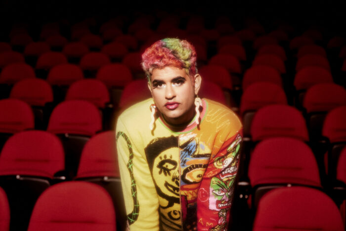 Photo of a person with short-multi-colored hair, wearing fake eyelashes and glamorous makeup in a theater of red seats.