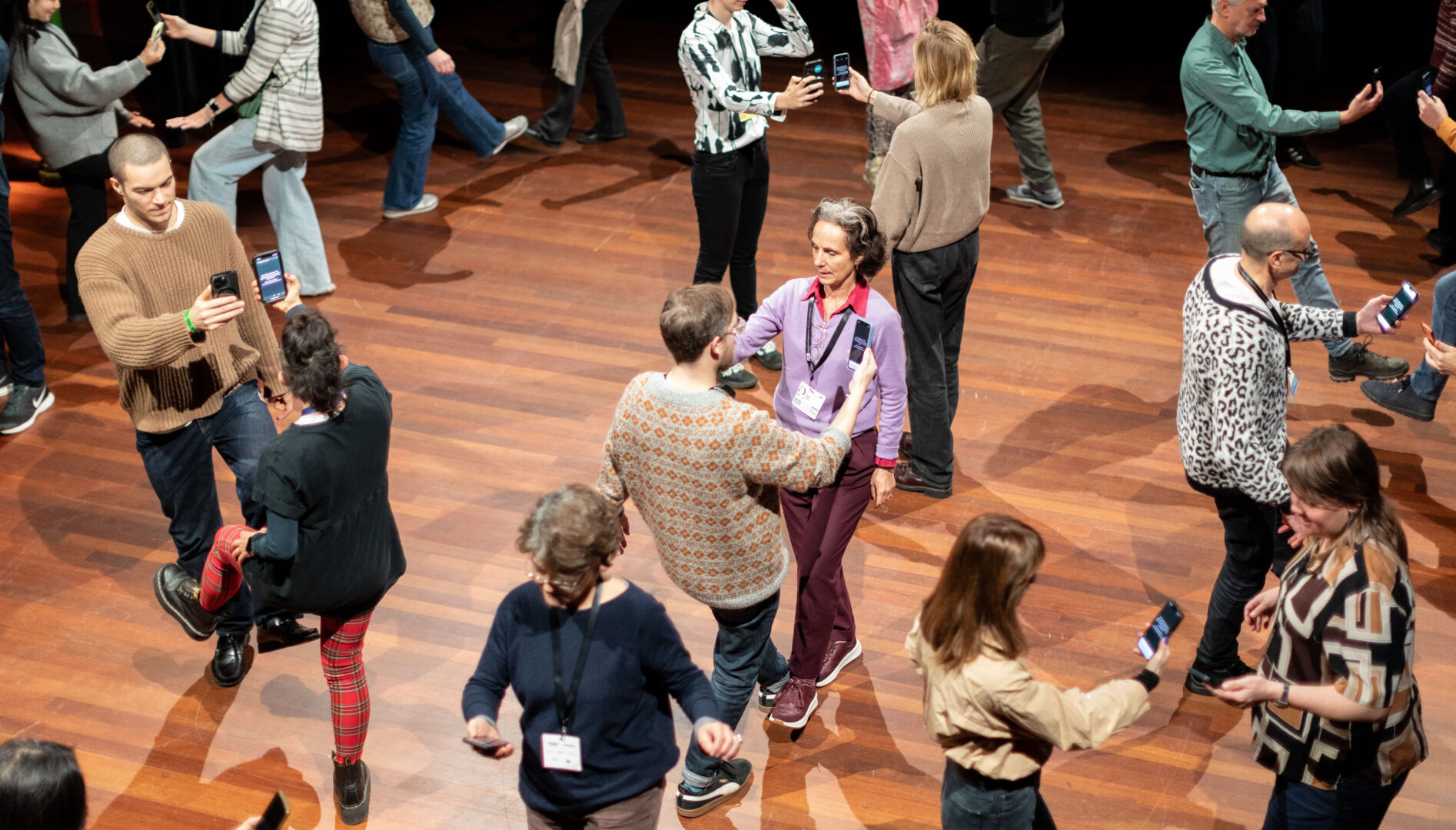 A crowd of people looking at their smartphones in pairs on a wooden floor.