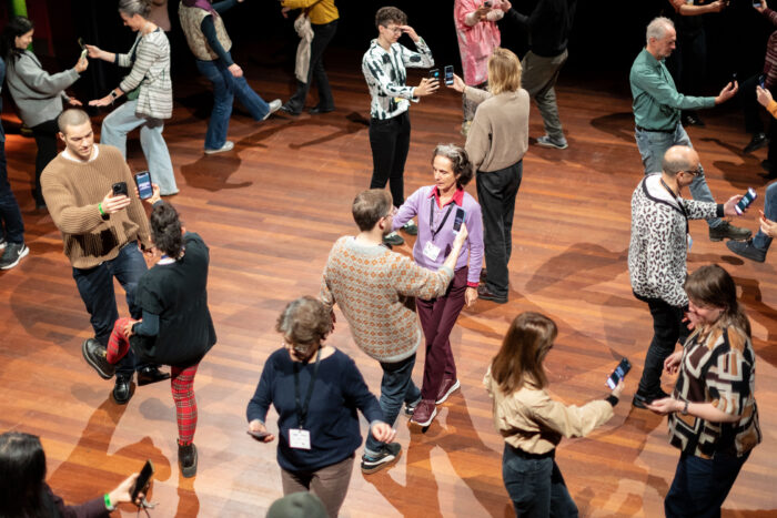 A crowd of people looking at their smartphones in pairs on a wooden floor.