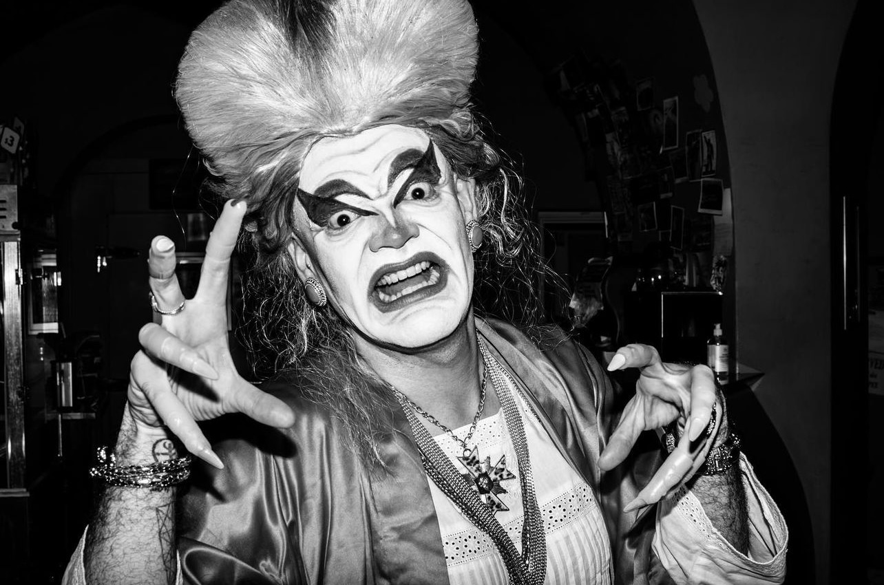 Black and white photo of a drag queen (Carla Rossi) wearing a big white wig and long, talon-like nails, making a scary face at the camera.