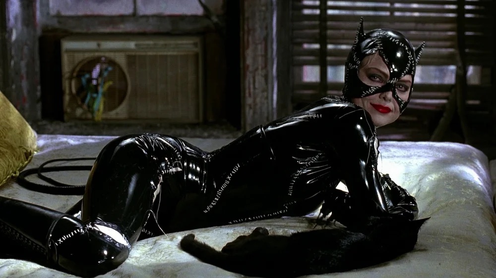 Film still of a woman in a shiny catsuit, laying on a bed next to a black cat.