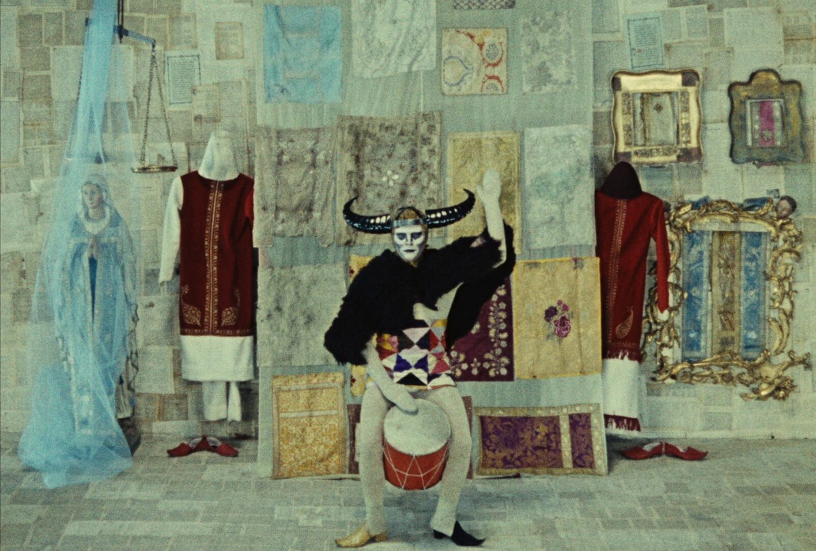 Film still of a surreal scene with mannequins and a Virgin mary statue at the back and a jester-like figure playing a drum in the foreground.
