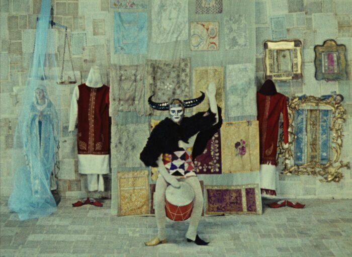 Film still of a surreal scene with mannequins and a Virgin mary statue at the back and a jester-like figure playing a drum in the foreground.