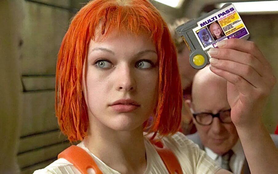 Film still of a white woman with an orange-red dyed bob haircut, holding up an ID card.