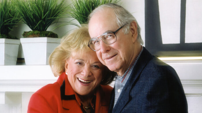 A smiling older white woman with blond hair, wearing a red jacket, standing next to a smiling older white man with eyeglasses and a blue suit coat on.