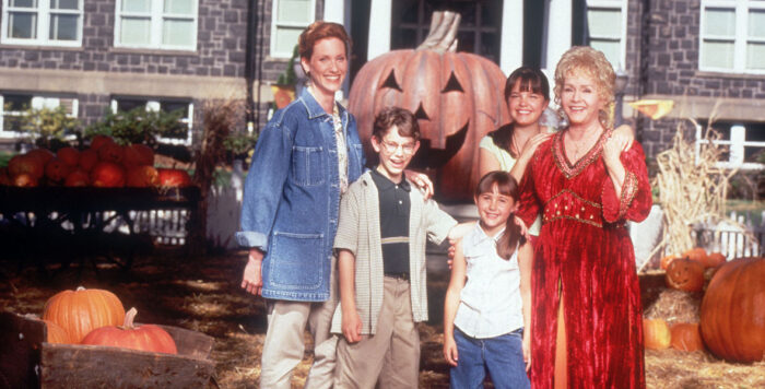 An multigenerational group of white people standing outside in an autumnal setting, with a big pumpkin behind them.