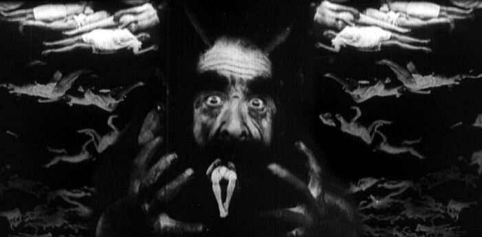 Black and white film still of a scary face in the middle and ghostlike figures around it.