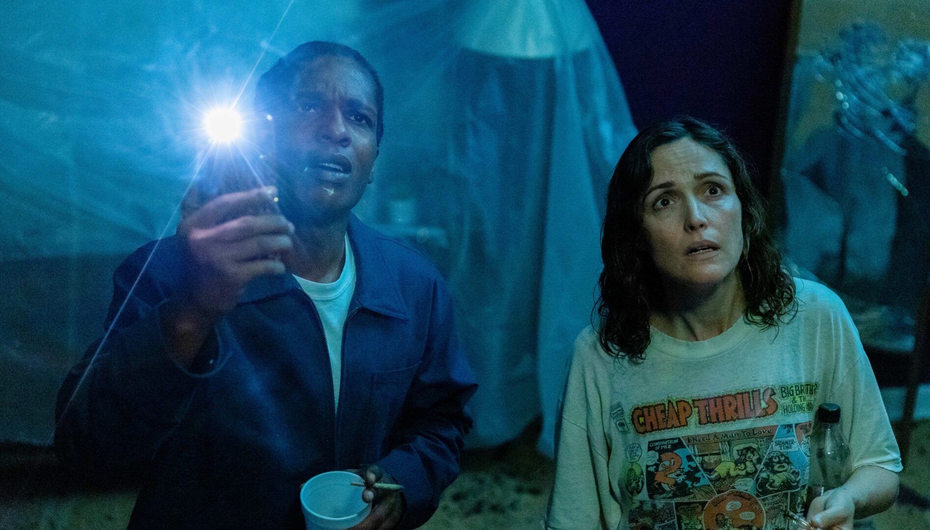 A film still of a nighttime scene of a Black man holding a cup in one hand and a holding up a phone with flashlight on in the other, standing next to a white woman with wet hair and a Cheap Thrills t-shirt on.